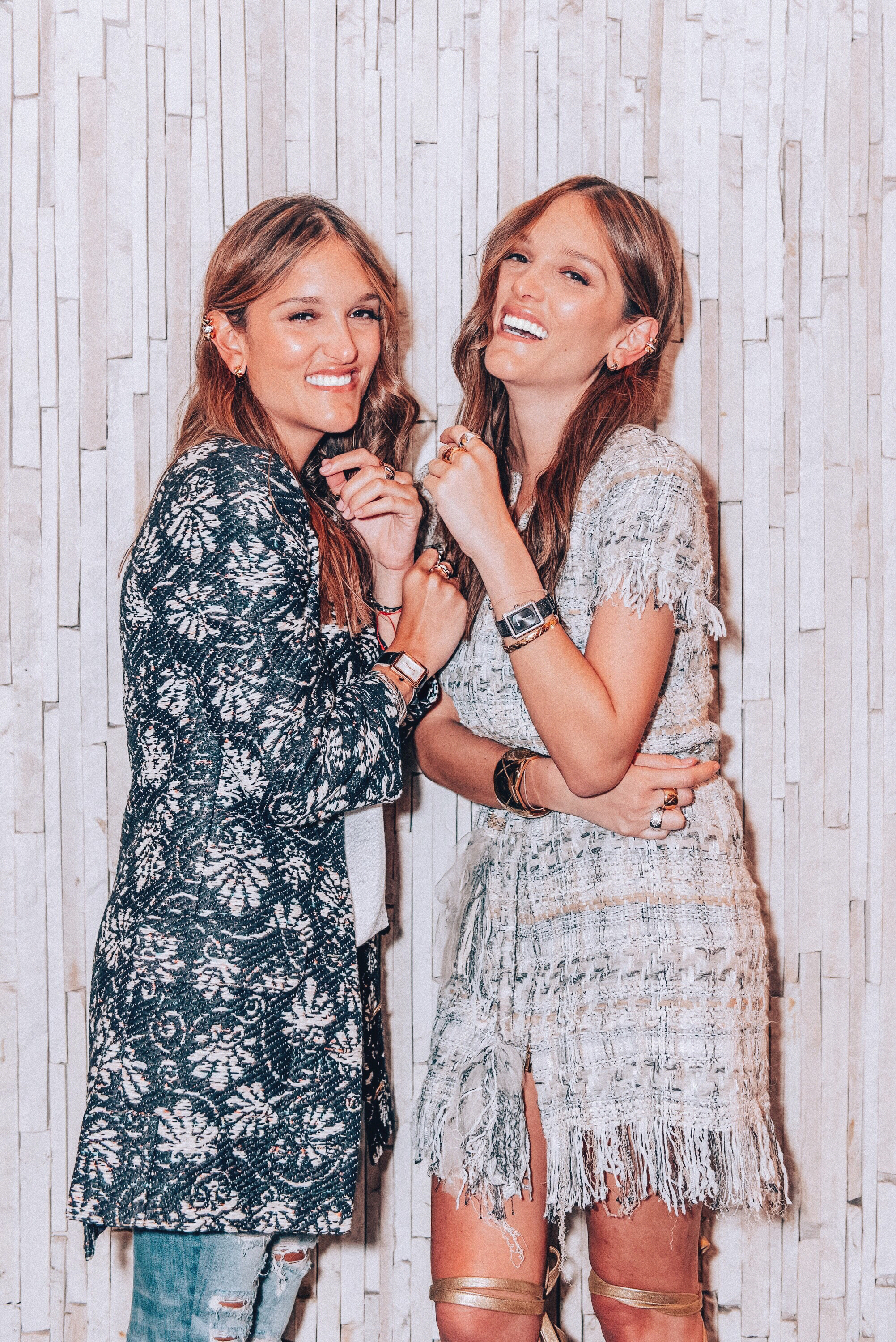 Two women wearing jewelry smile together against a pale tiled wall